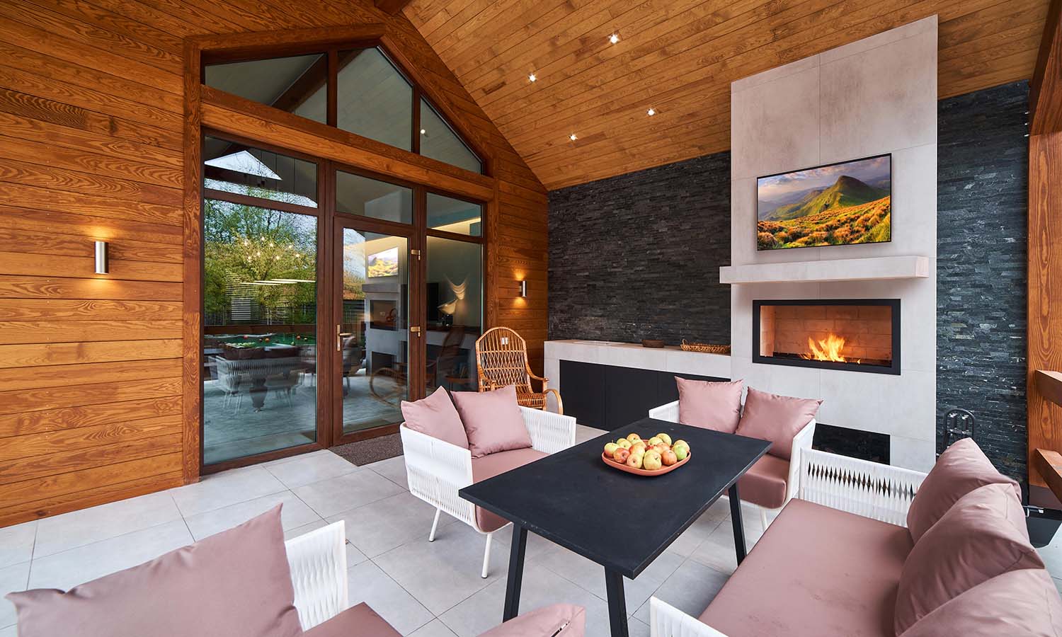 A stylish outdoor living space with a modern fireplace, mounted TV displaying a scenic landscape, comfortable seating, and a dining table with fresh fruit, all under a wood-paneled ceiling.
