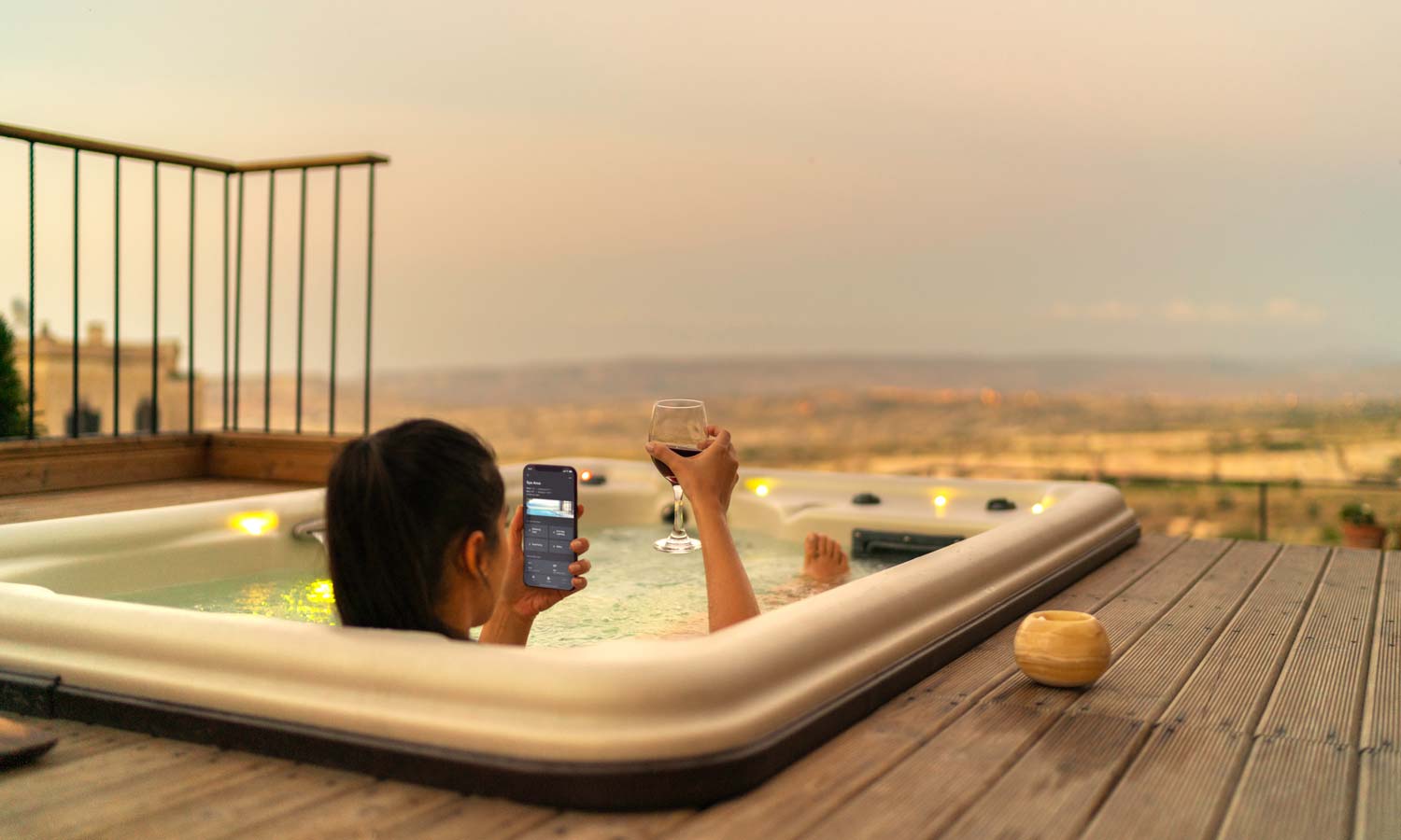 A woman relaxing in an outdoor hot tub at sunset, holding a glass of wine in one hand and a smartphone displaying a smart home control app in the other, overlooking a scenic landscape.